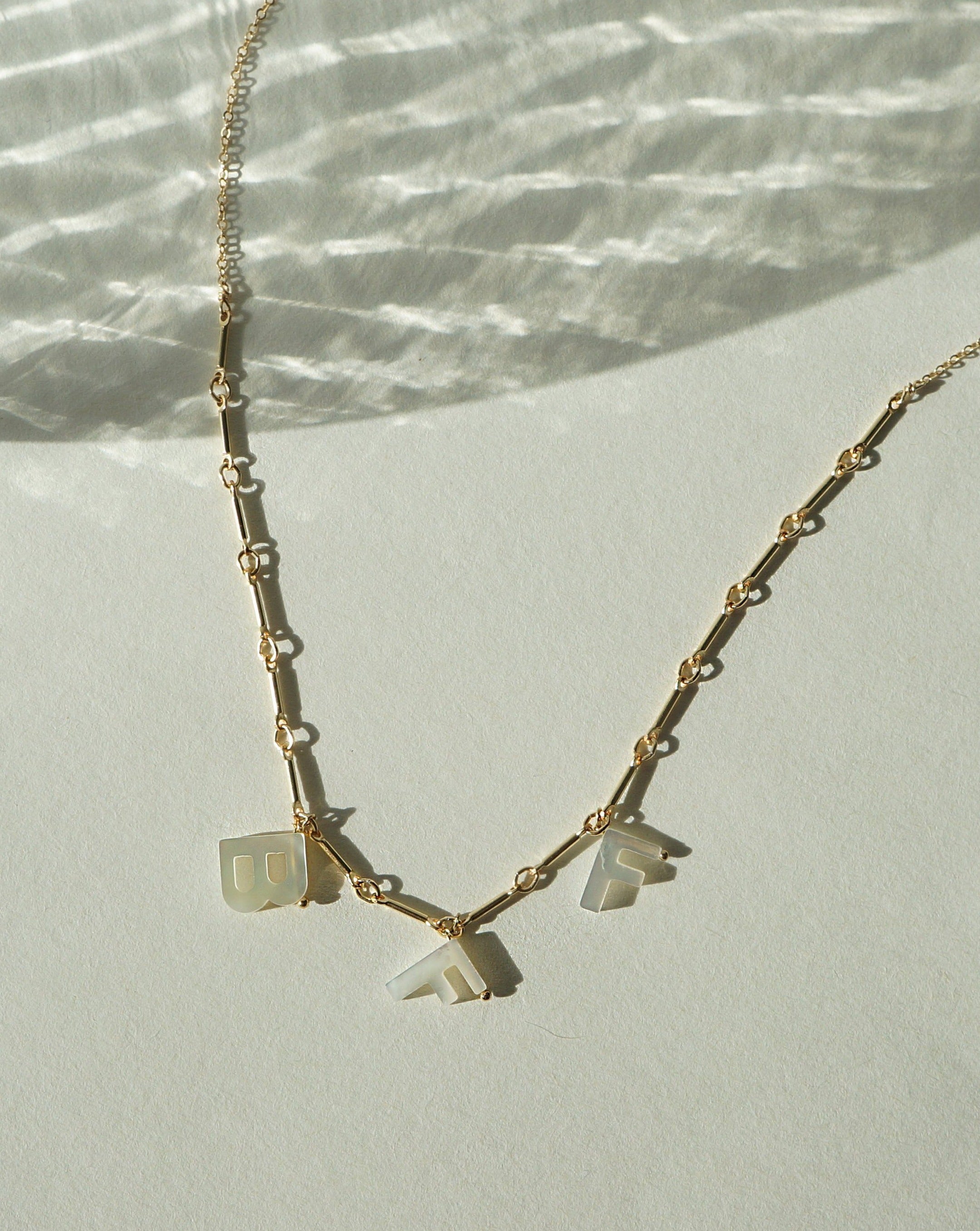 Mama Necklace by KOZAKH. A 16 to 18 inch adjustable length necklace with linked bar chain on bottom half and flat link chain on top half, crafted in 14K Gold Filled, featuring a customizable word made from hand carved Mother of Pearl letters.