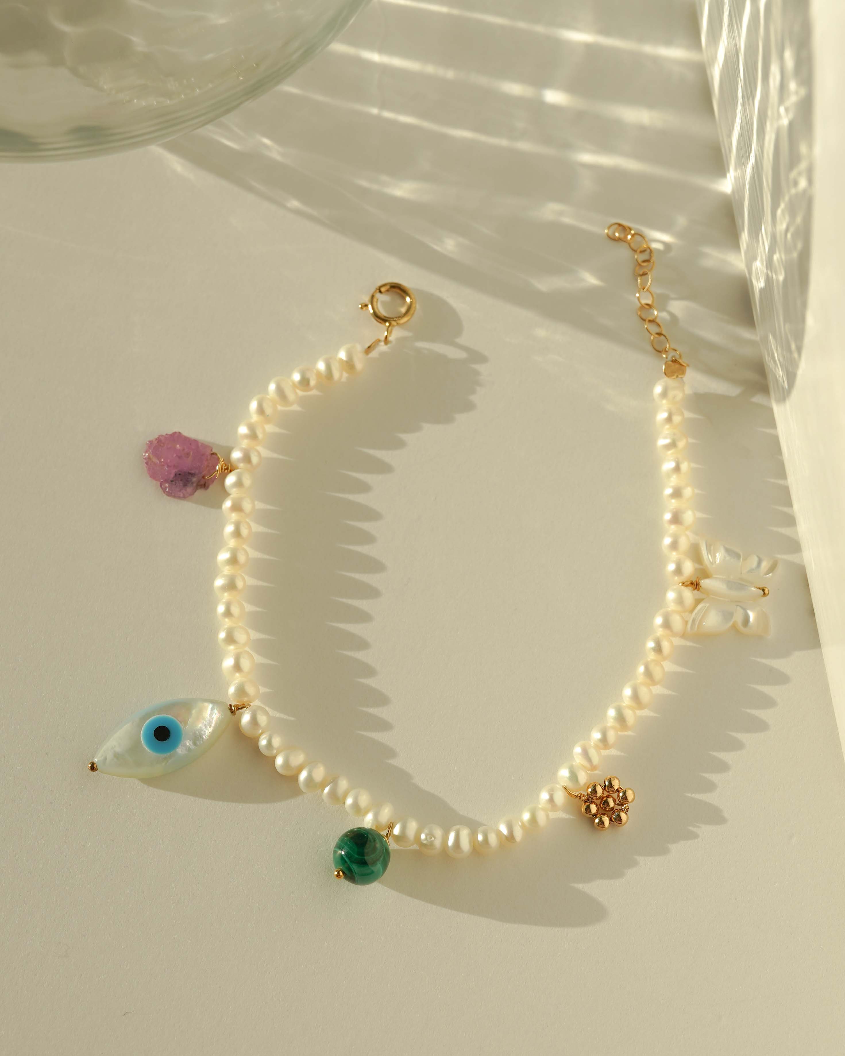 Rylan Bracelet by KOZAKH. A 6 to 7 inch adjustable length, 3mm freshwater pearl strand bracelet, crafted in 14K Gold Filled, featuring a Pink Tourmaline slice, a Mother of Pearl butterfly charm, a round Malachite gemstone, and Mother of Pearl evil eye charm.
