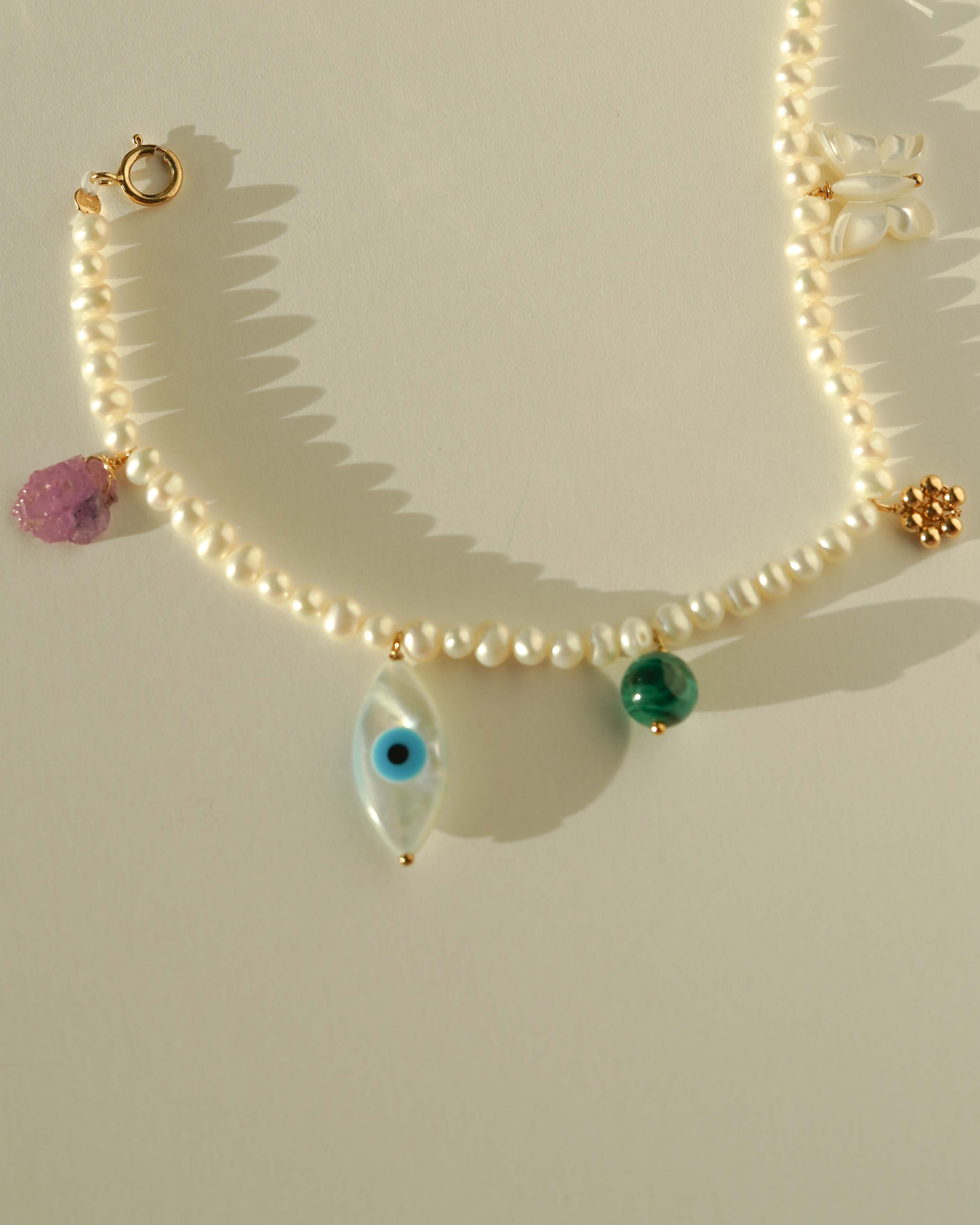 Rylan Bracelet by KOZAKH. A 6 to 7 inch adjustable length, 3mm freshwater pearl strand bracelet, crafted in 14K Gold Filled, featuring a Pink Tourmaline slice, a Mother of Pearl butterfly charm, a round Malachite gemstone, and Mother of Pearl evil eye charm. 
