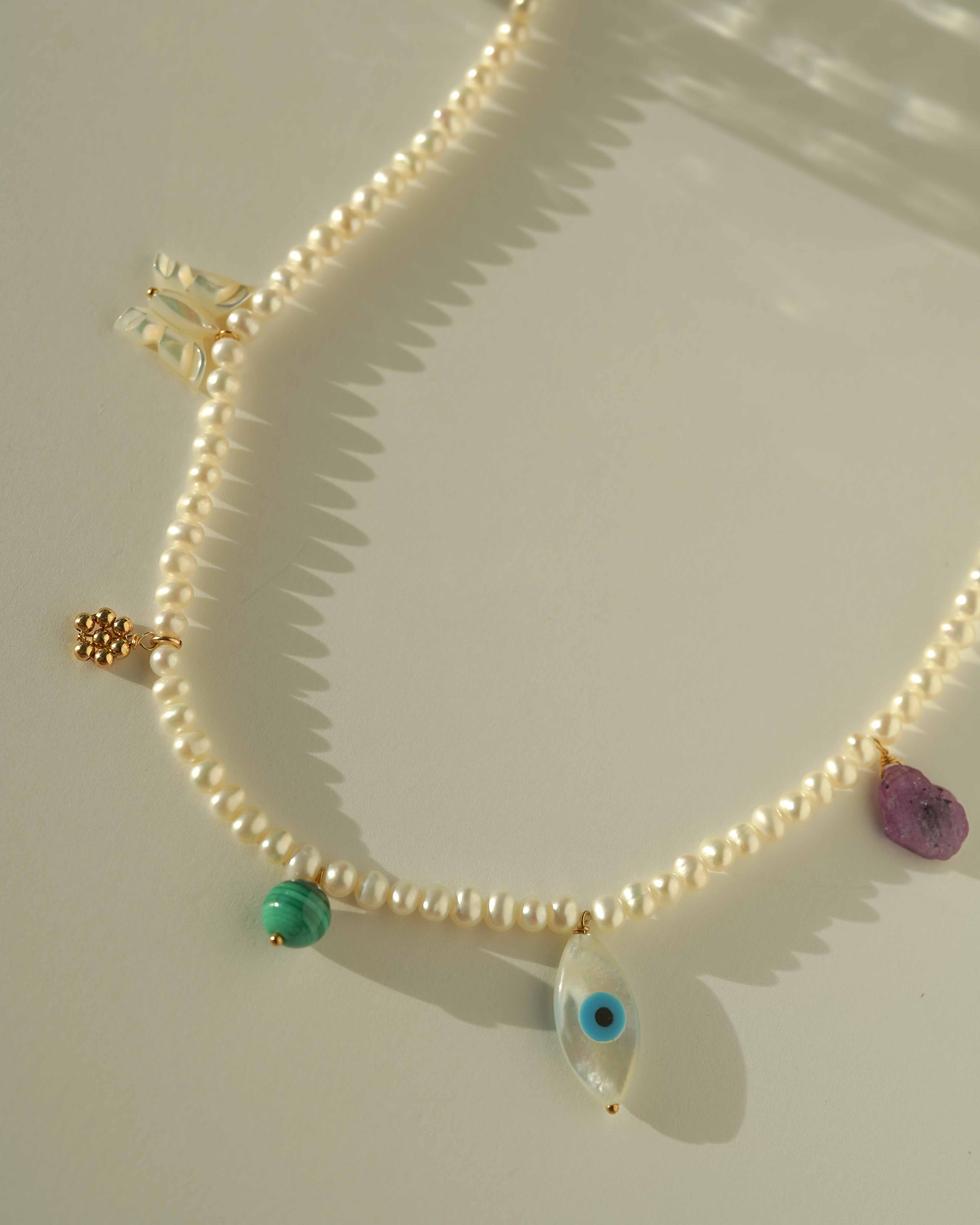 Valery Necklace by KOZAKH. A 16 to 18 inch adjustable length necklace, with 16 inch strand of 3mm Freshwater Pearls, clasp crafted in 14K Gold Filled, featuring Pink Tourmaline slice, Mother of Pearl butterfly charm, smooth round Malachite gemstone, Mother of Pearl eye charm, and handmade gold beaded daisy charm.
