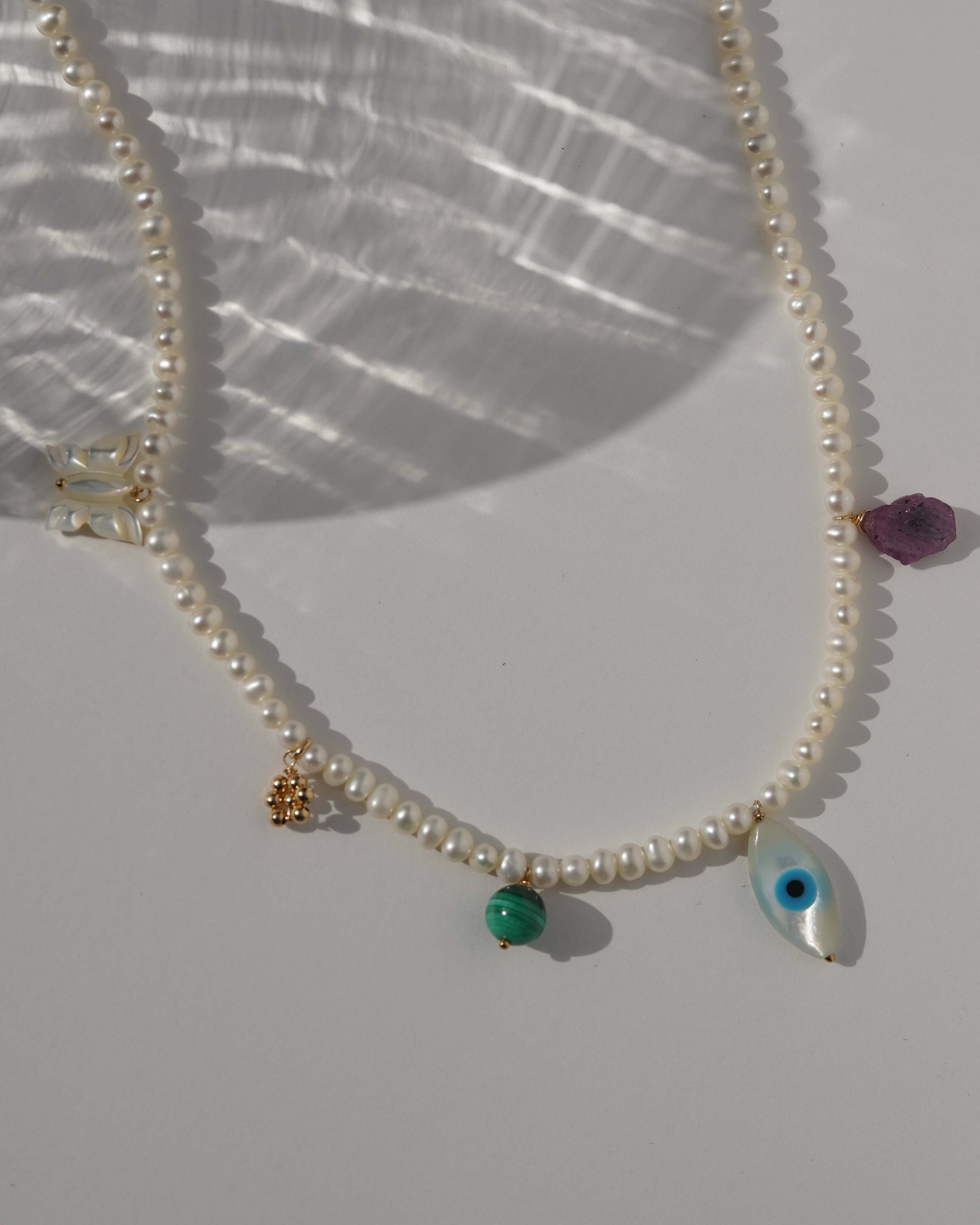 Valery Necklace by KOZAKH. A 16 to 18 inch adjustable length necklace, with 16 inch strand of 3mm Freshwater Pearls, clasp crafted in 14K Gold Filled, featuring Pink Tourmaline slice, Mother of Pearl butterfly charm, smooth round Malachite gemstone, Mother of Pearl eye charm, and handmade gold beaded daisy charm.