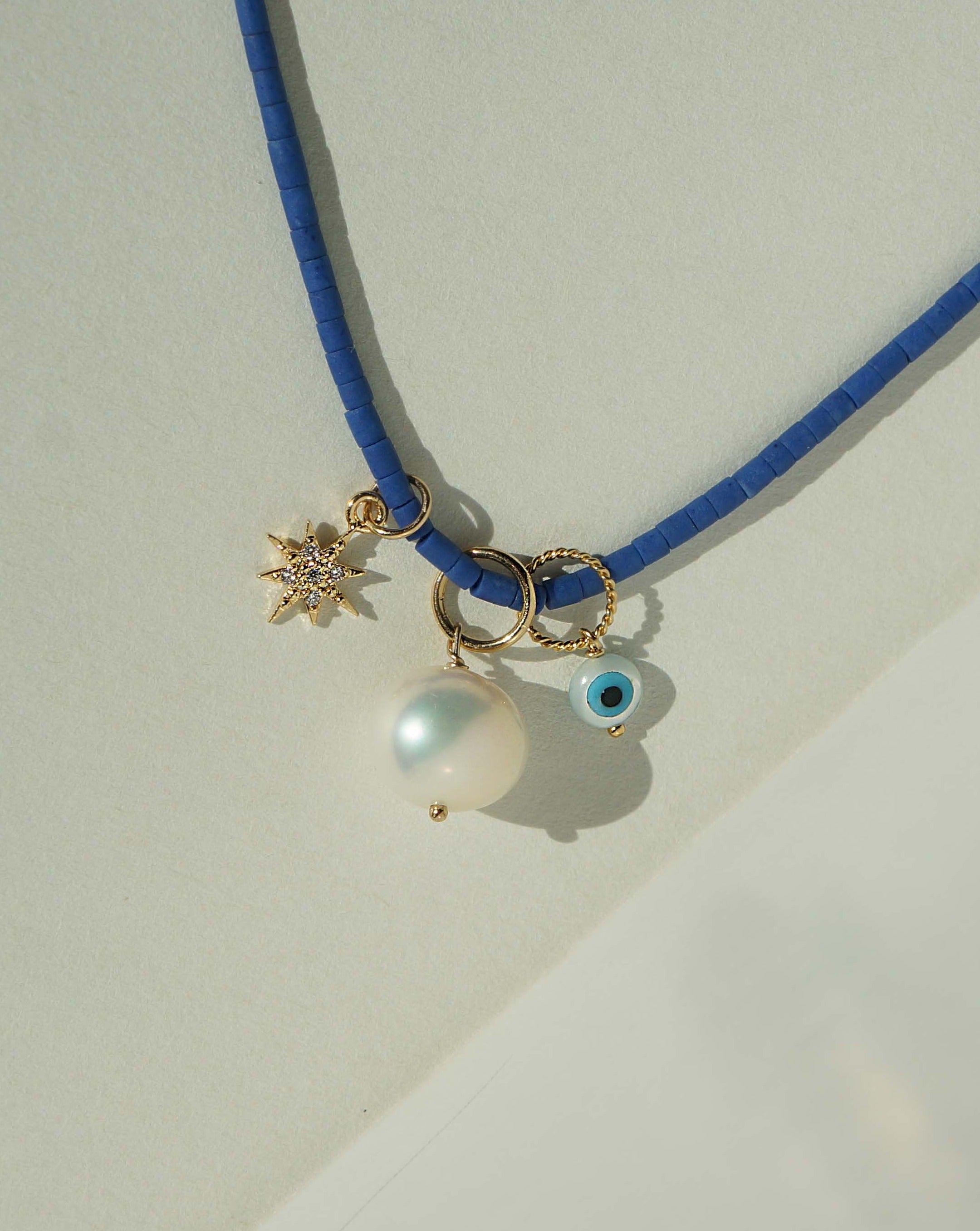 Brook Dark Blue Necklace by KOZAKH. A 13 to 16 inch adjustable length colored Afghan Beads strand necklace in 14K Gold Filled, featuring a hand carved Mother of Pearl evil eye charm, a round Freshwater Pearl, and a Cubic Zirconia encrusted star charm.