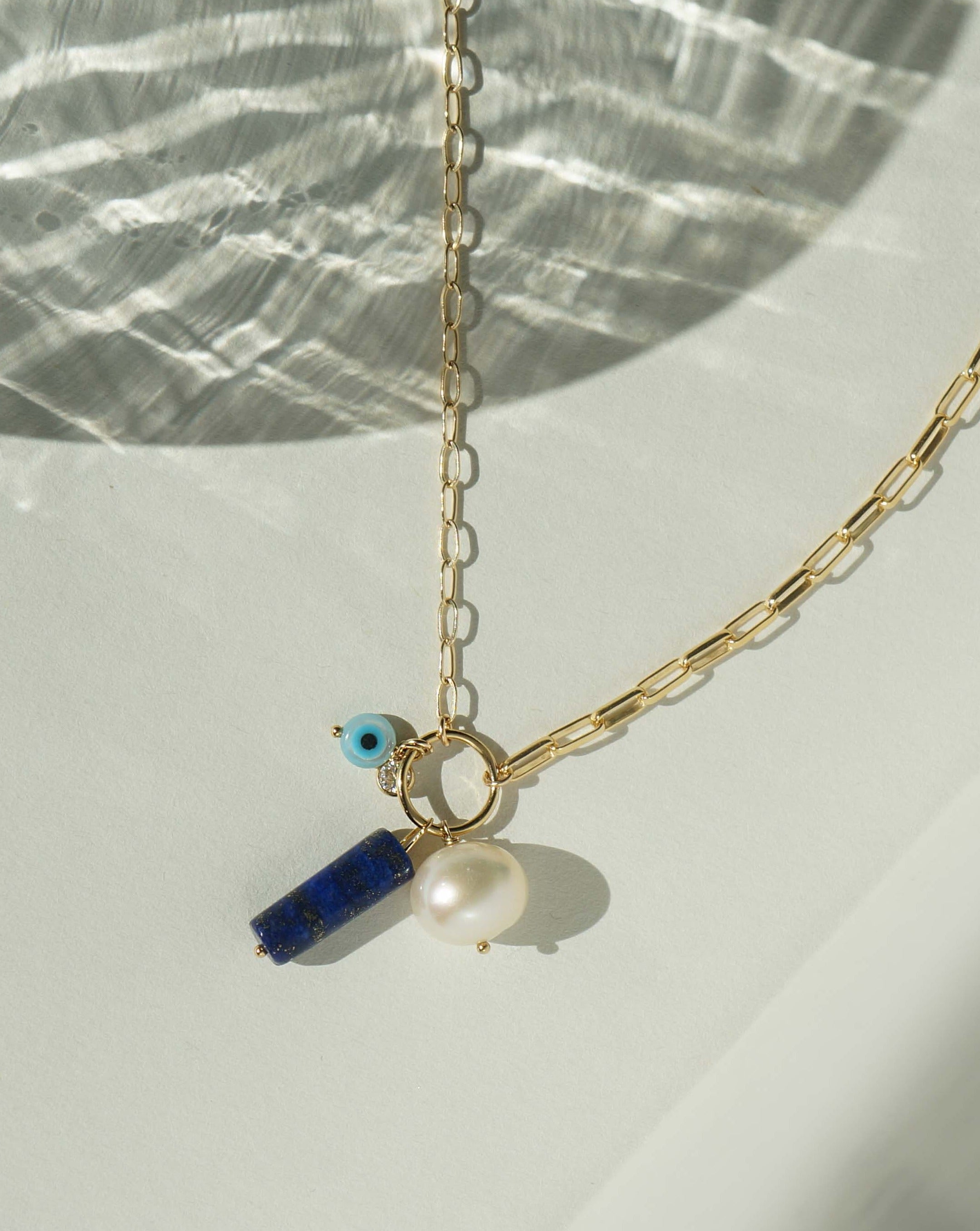 Lucky Necklace by KOZAKH. A 16 to 18 inch adjustable length necklace with flat link chain on one half and mini paperclip chain on the other half, crafted in 14K Gold Filled, featuring a cylindrical Lapis, a round Freshwater Pearl, and a hand carved Mother of Pearl Evil Eye.