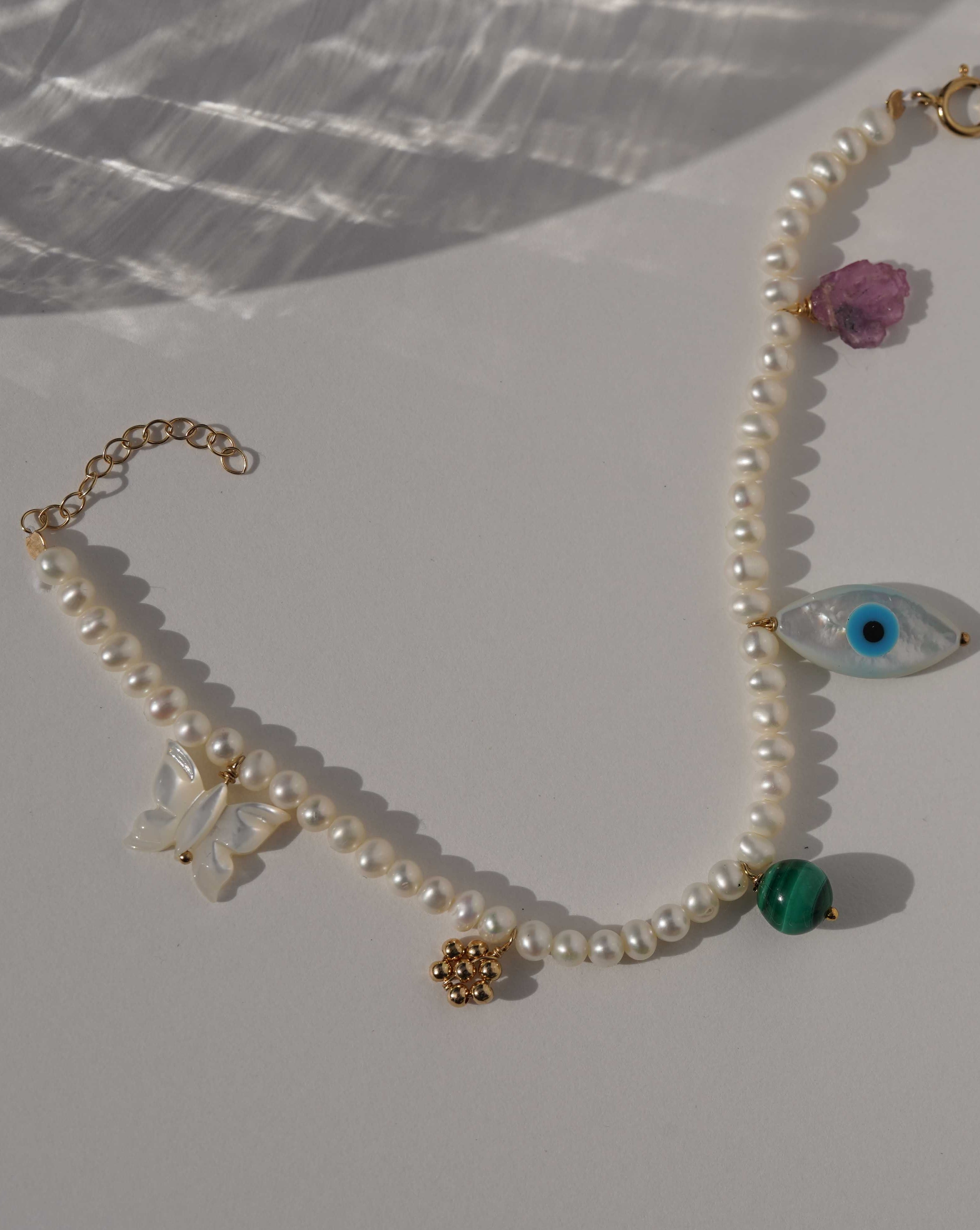 Rylan Bracelet by KOZAKH. A 6 to 7 inch adjustable length, 3mm freshwater pearl strand bracelet, crafted in 14K Gold Filled, featuring a Pink Tourmaline slice, a Mother of Pearl butterfly charm, a round Malachite gemstone, and Mother of Pearl evil eye charm. 